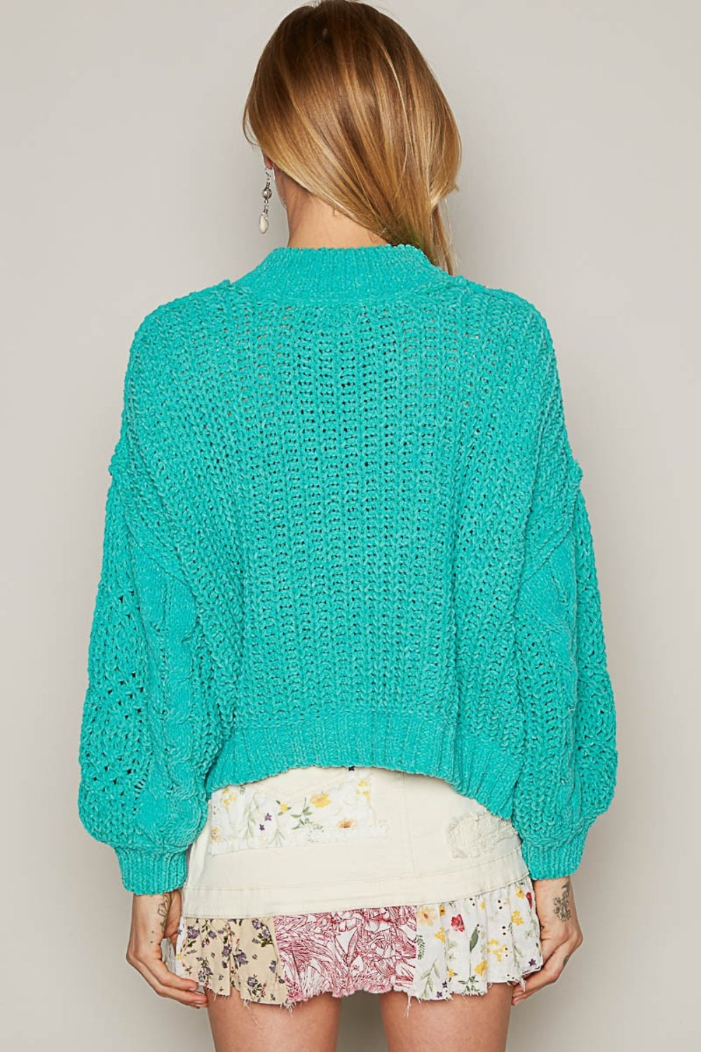 A person in a turquoise POL Mock Neck Cable Knit Sweater with a textured pattern is sitting outdoors.