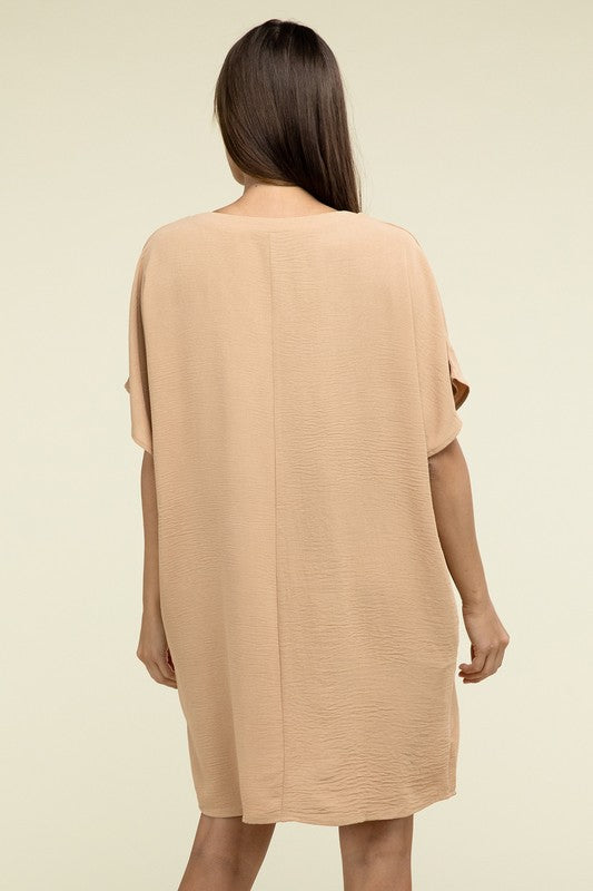 A woman wearing a loose light brown Woven Airflow V Neck T-Shirt Dress with Pockets stands against a plain background. Made from breathable fabric, this summer dress includes pockets where her hands rest as she smiles slightly.