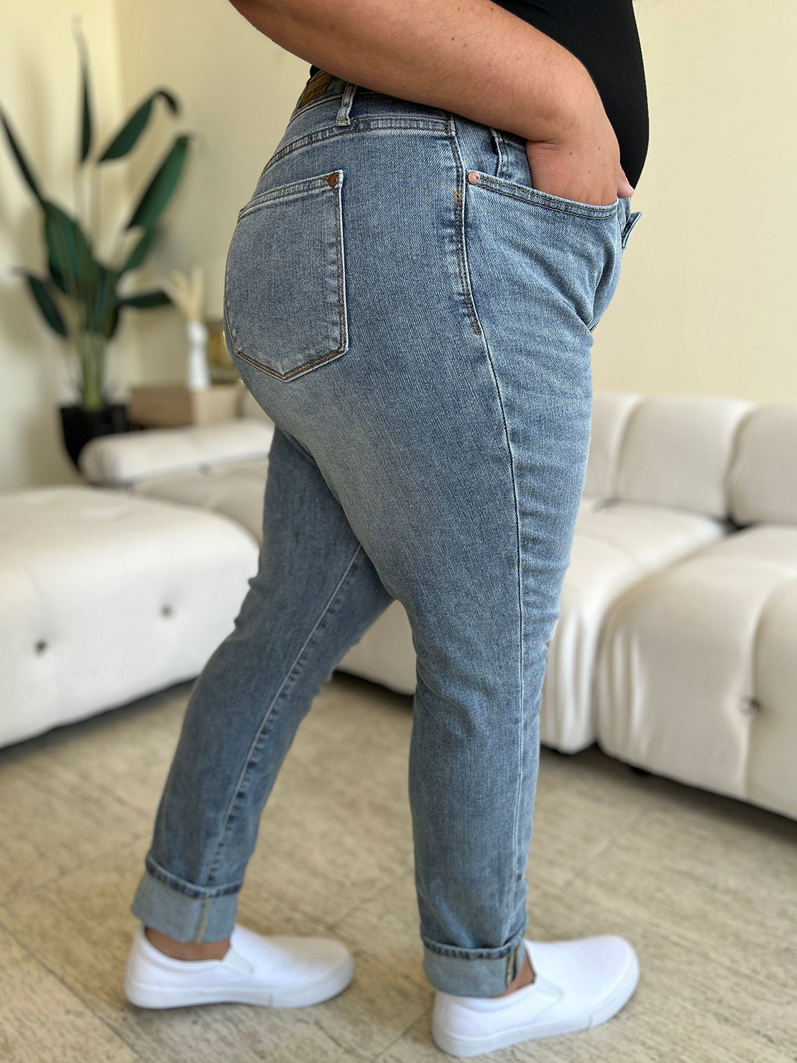 A person wearing Judy Blue Full Size High Waist Cuff Hem Jeans and white slip-on shoes stands in a living room with a white couch and plants in the background.