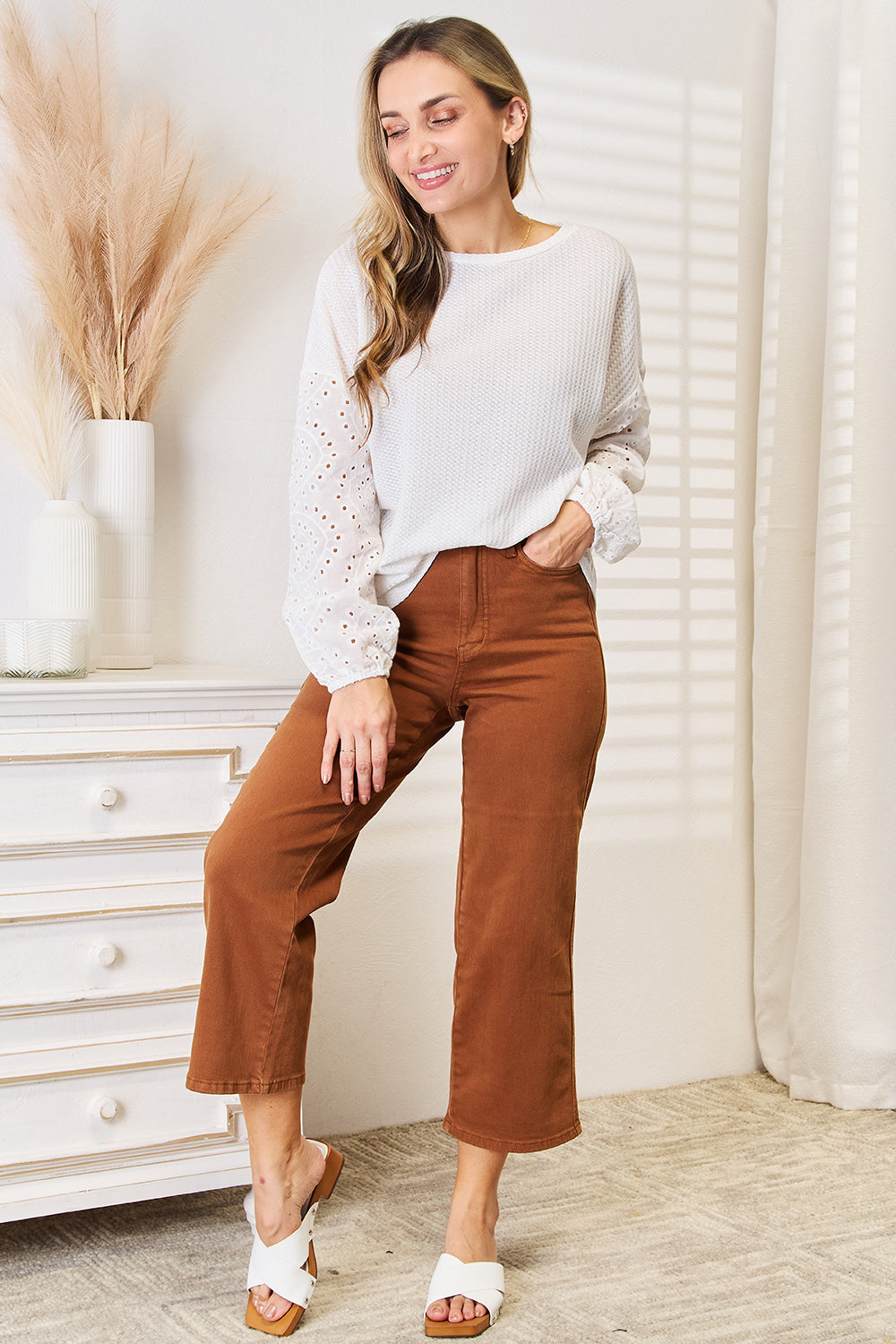 A woman wearing a Shiny Eyelet Dropped Shoulder Round Neck Blouse and brown pants stands in a room with sunlight streaming through blinds. She is smiling and has her left hand on her hip.