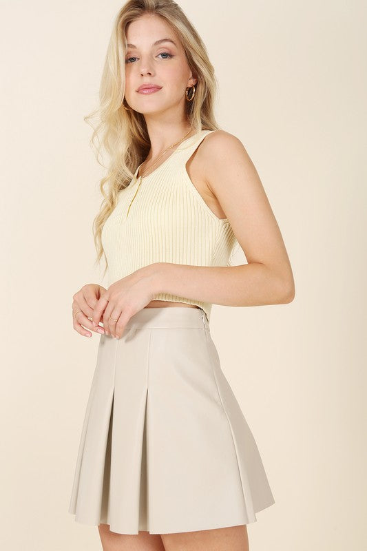 A woman in a beige tank top stands against a light background, looking to her right while wearing the Vegan Leather Pleated Mini Skirt featuring a side zipper.