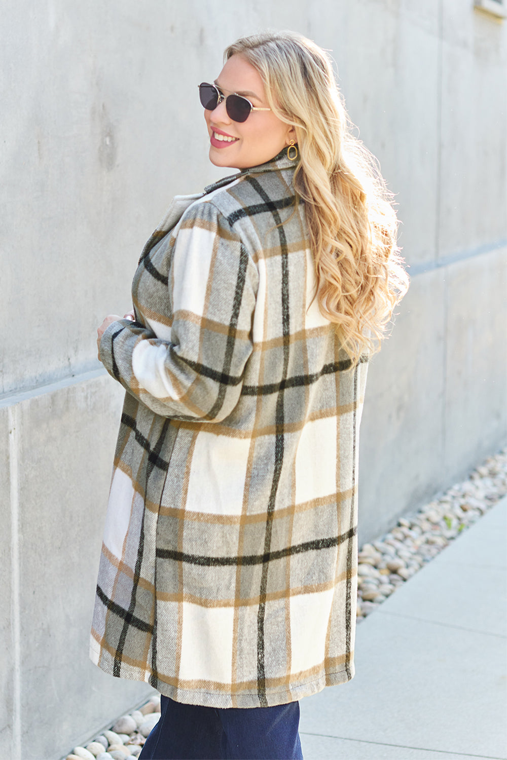 A woman stands on a sidewalk wearing the Double Take Full Size Plaid Button Up Lapel Collar Coat in pink and white over a white top and blue jeans, with her right hand lightly touching her hair. The polyester fabric of the coat makes it durable and perfect for an easy machine wash cold.