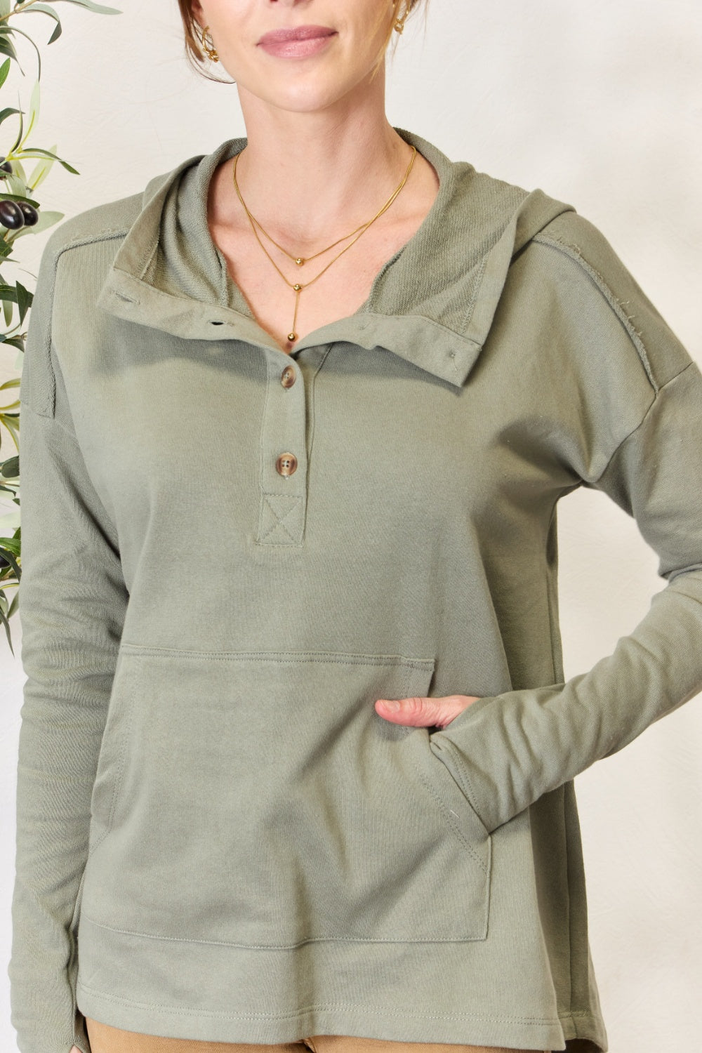 A person stands with hands in the pockets of their light green Culture Code Full Size Half Button Hoodie and brown pants. A plant is visible on the left side of the image.