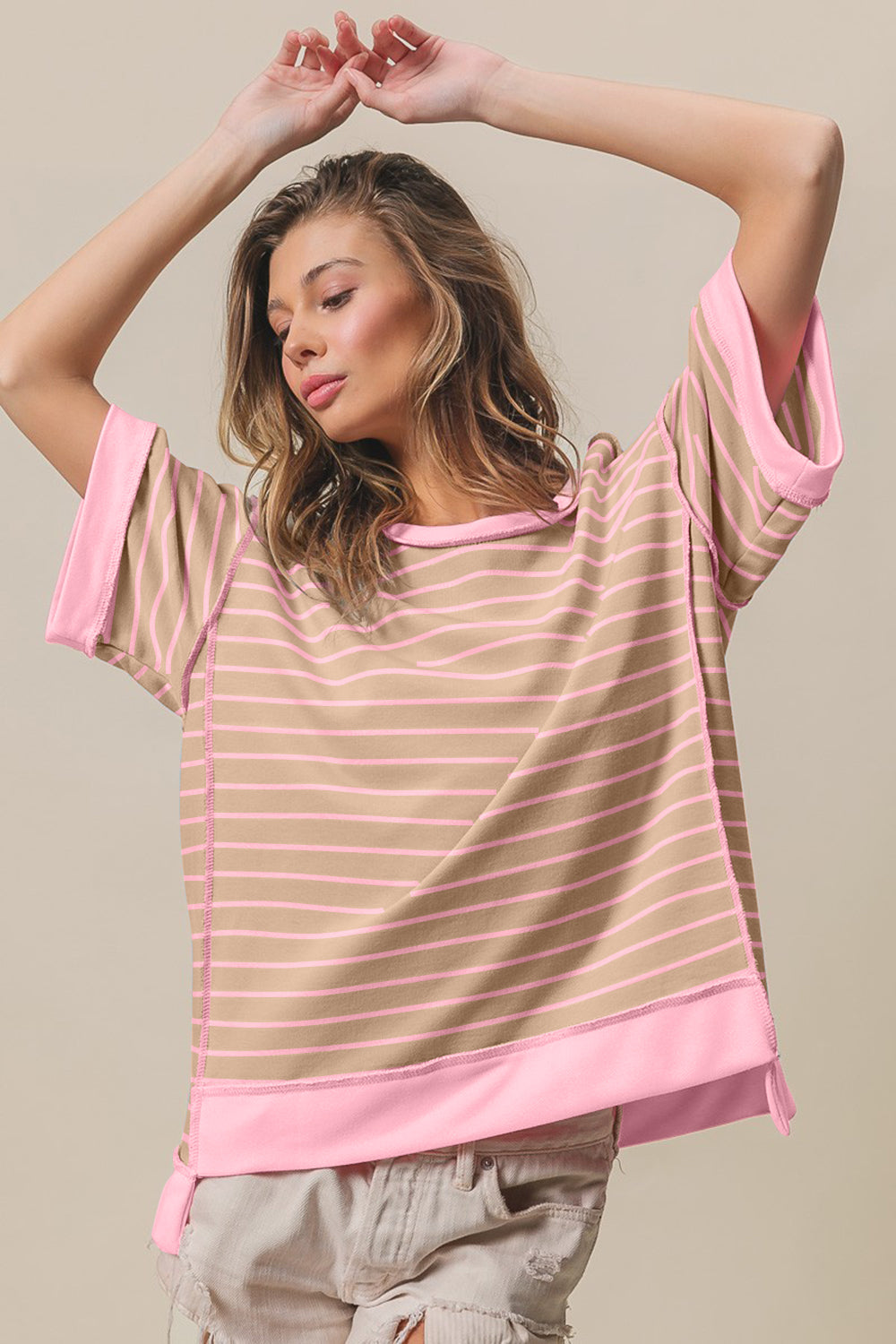 A person wearing a wide-brimmed hat, an oversized BiBi Exposed Seam Stripe Contrast T-Shirt, and distressed denim shorts stands with one arm raised and eyes closed against a beige background.