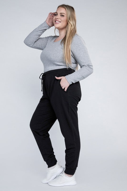 A person stands against a white background, wearing a white t-shirt, grey Plus-Size Jogger Pants with an adjustable waistband, and white sneakers. They have long blonde hair and are posing with one hand on their hip.