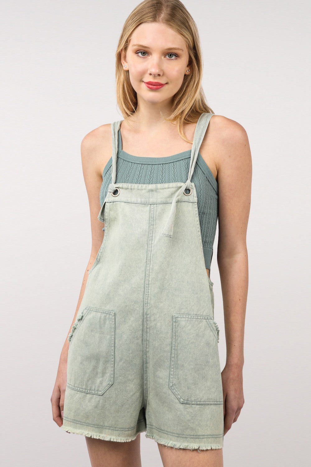 A young woman with shoulder-length hair wearing a green knit top and the VERY J Washed Frayed Hem Denim Overall stands against a plain background, effortlessly blending vintage fashion with a modern twist.