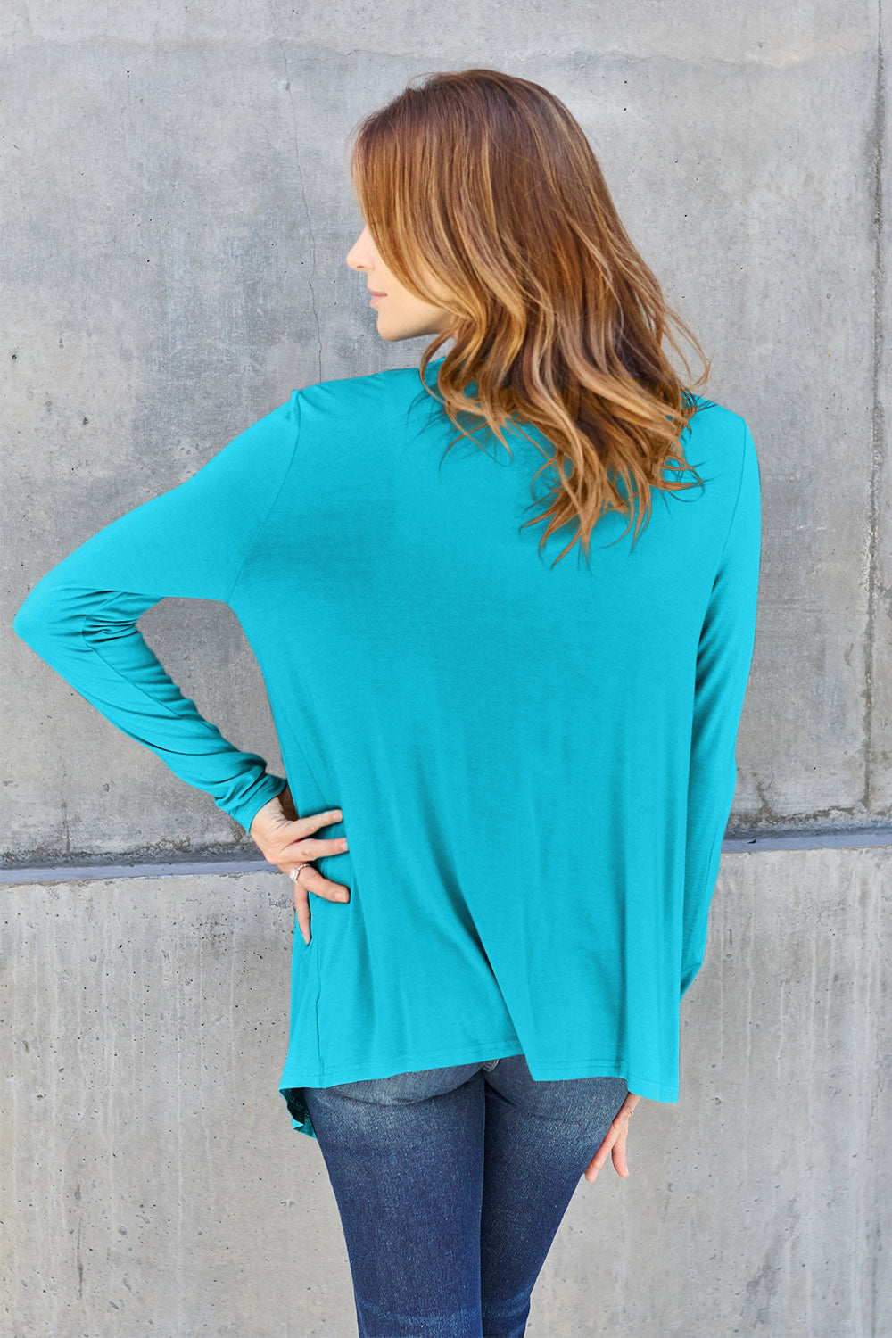 A woman wearing the Basic Bae Full Size Open Front Long Sleeve Cardigan in navy blue, paired with a white top and blue jeans, stands against a concrete wall. She touches her hair and gazes to the side, embodying a chic yet simple style. The slightly stretchy materials of the cardigan ensure comfort while maintaining its stylish simplicity. For easy care, machine wash it cold.