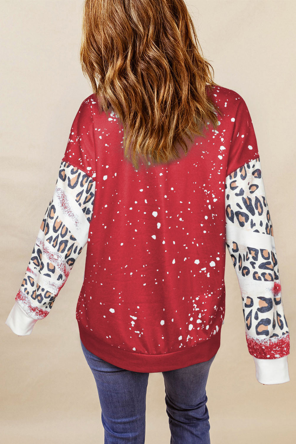 A person with wavy hair is seen from the back wearing the Fiery Red Tie Dye Leopard Drop Shoulder Sweatshirt, creating a stylish look with its red hue adorned with paint splatters and leopard print sleeves. They are also wearing jeans, showcasing a casual sweatshirt that's perfect for any laid-back occasion.