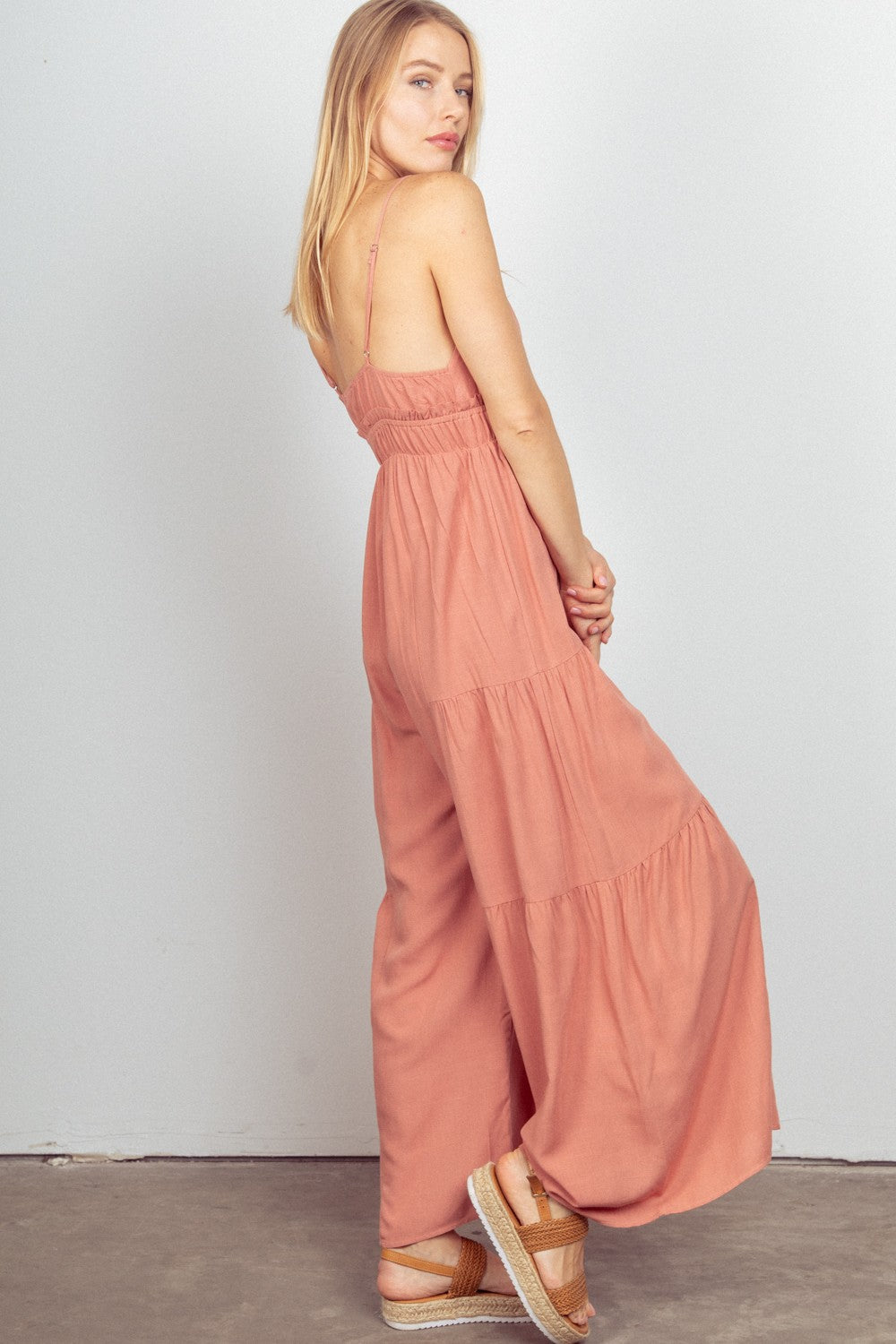 A woman stands against a plain background, wearing the VERY J Sleeveless Ruched Wide Leg Jumpsuit in pink, featuring an elastic waist and adjustable straps. She completes the look with crossover sandals.