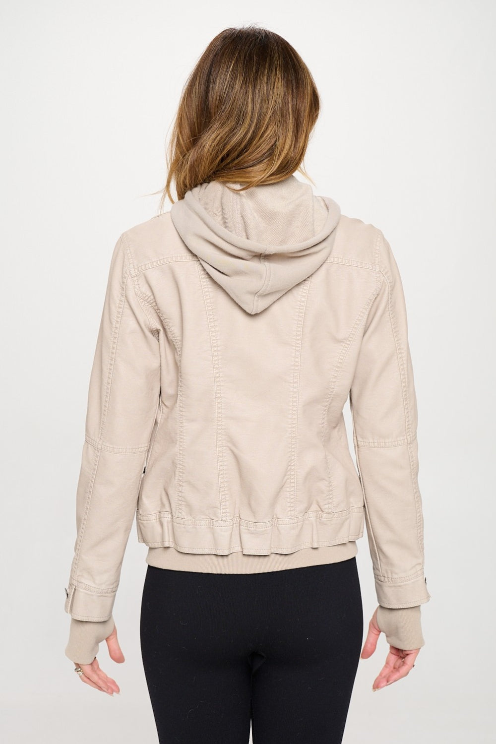 A woman wearing the Coalition LA Double Zip Hooded Long Sleeve Jacket in beige, made from vegan leather, stands against a plain white background, paired with a black top and leggings.