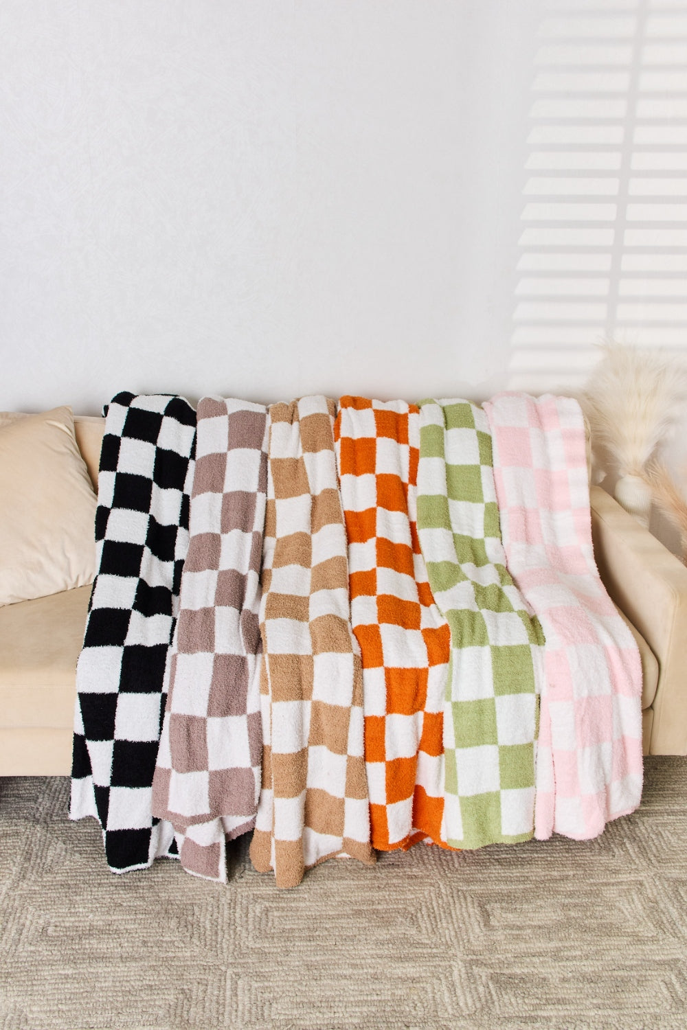 A person stands holding a large Cuddley Checkered Decorative Throw Blanket, smiling, in a room with a sofa and a potted plant. The blanket features a black and white checkerboard pattern, is made from 100% polyester, and imported for quality.