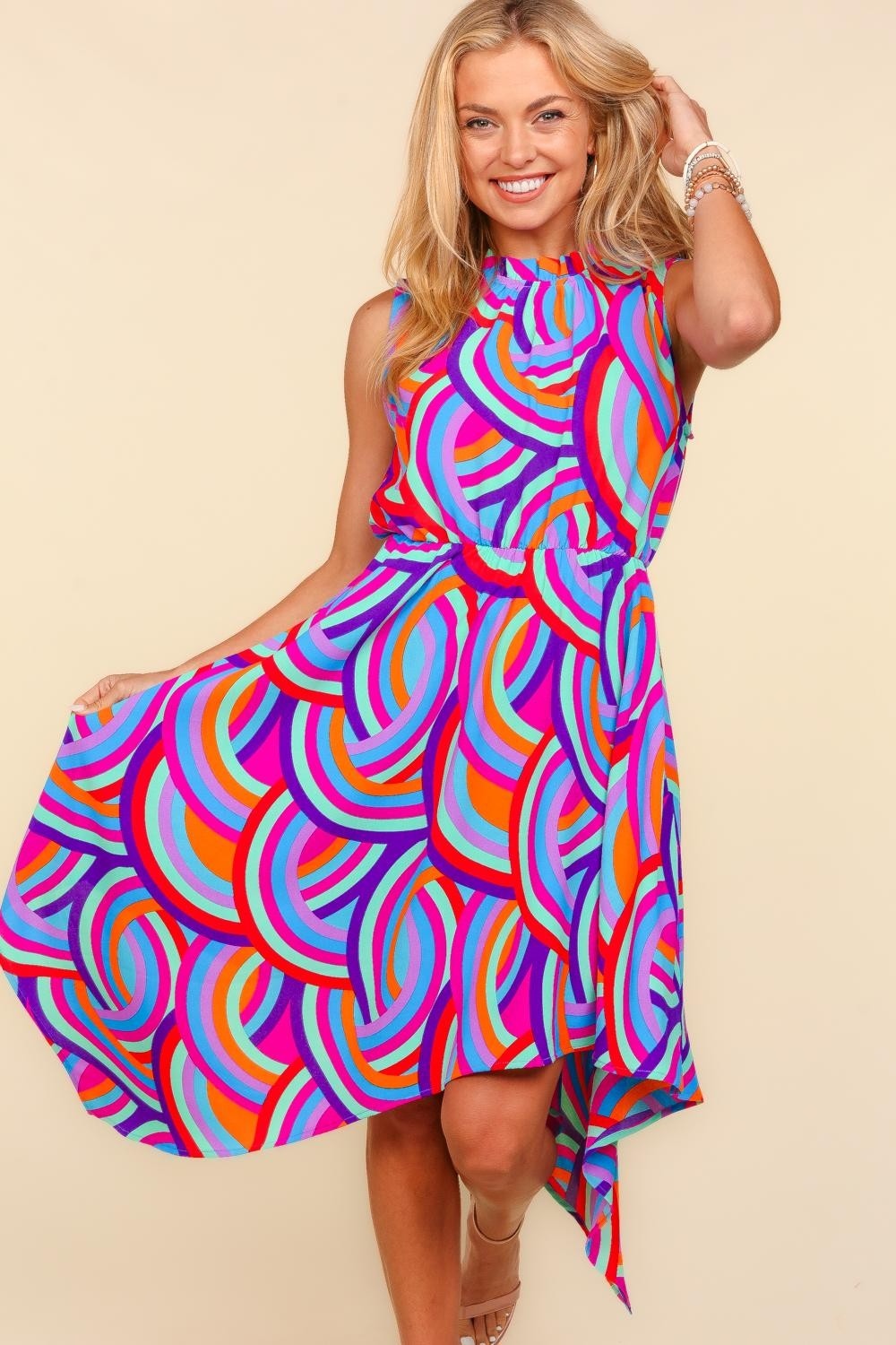 Against a beige backdrop, an individual models the Haptics Full Size Mock Neck Sleeveless Printed Dress, which features an eye-catching asymmetrical pattern. The lively blend of pink, blue, orange, and purple swirls crafts a captivating visual symphony.