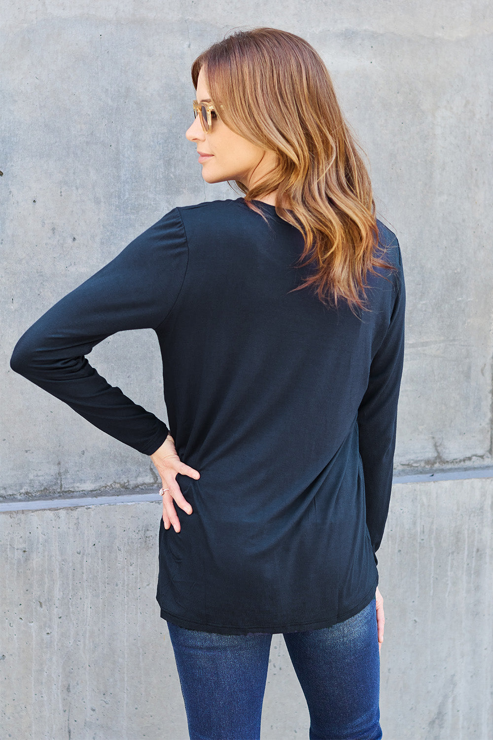 A woman with long hair wearing sunglasses, a Basic Bae Full Size Round Neck Long Sleeve Top in turquoise, and jeans is standing in front of a concrete wall with one hand on her hip.