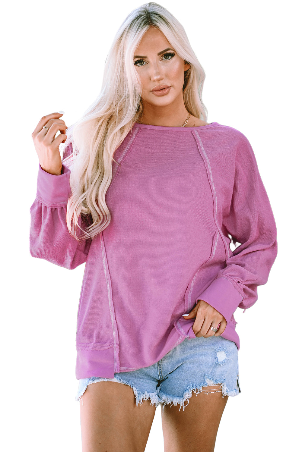 A person with long blonde hair is seen from the back wearing a Pink Exposed Seam Round Neck Terry Pullover and distressed denim shorts. The trendy look complements the casual outfit as they stand outdoors in front of a building with a window and a lantern.