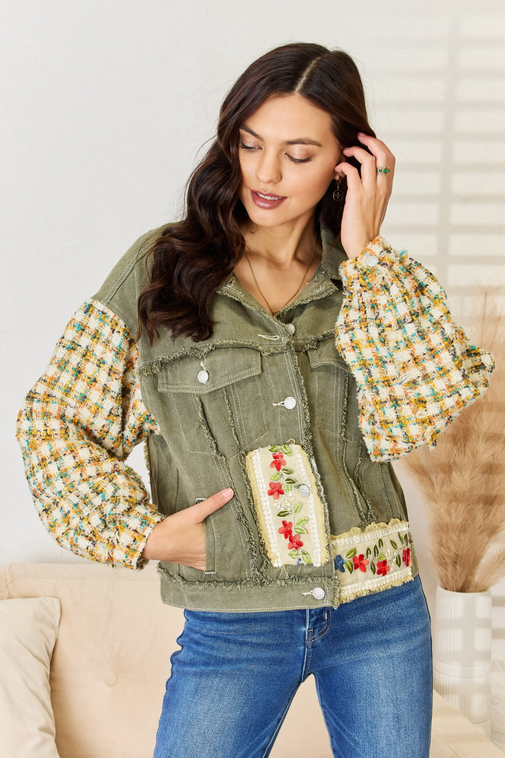 A woman with long hair stands indoors, wearing the POL Embroidered Button Down Raw Hem Shacket with floral designs and contrast tweed sleeves, paired with blue jeans. She has one hand touching her hair and the other resting in her jacket pocket.