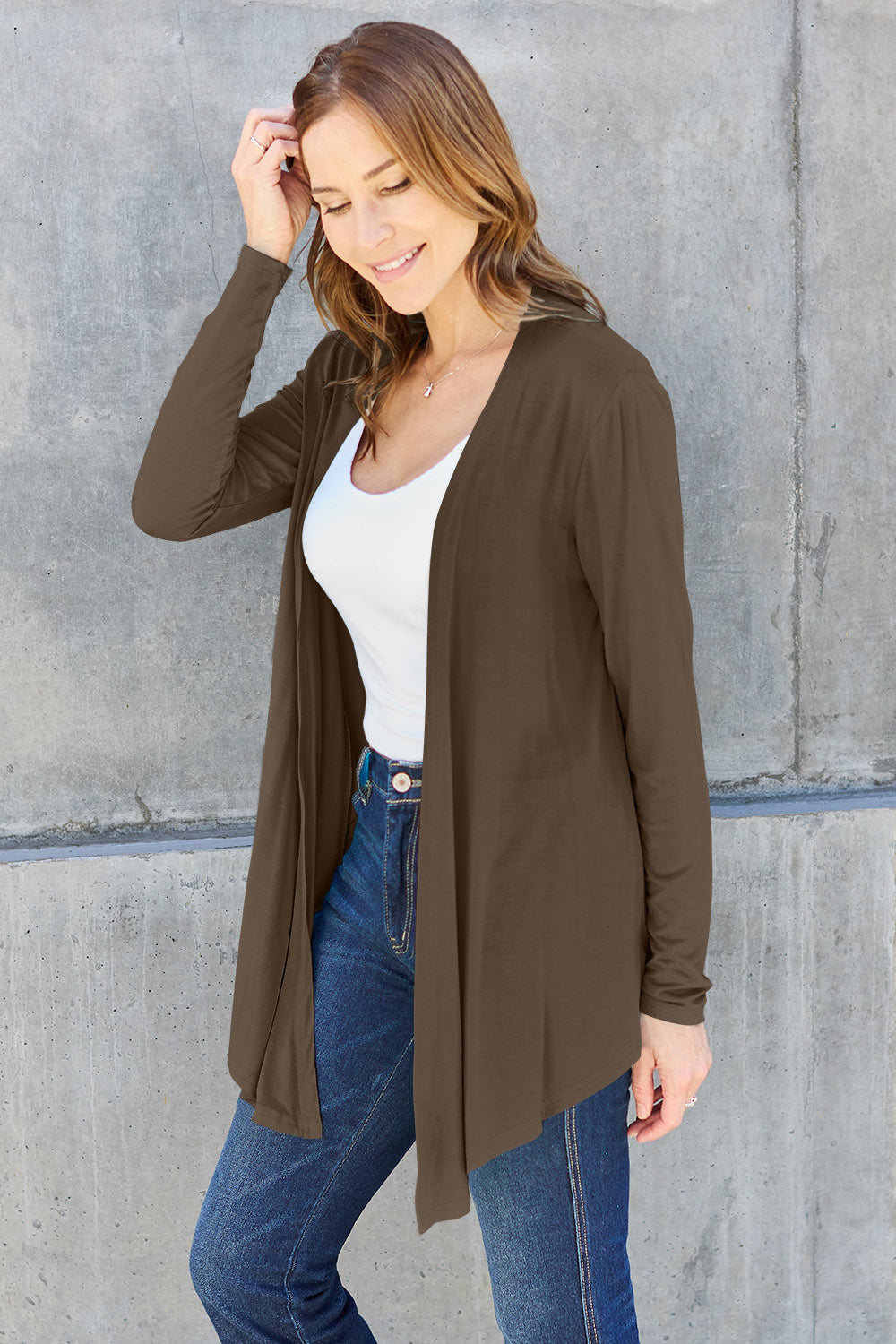 A woman wearing the Basic Bae Full Size Open Front Long Sleeve Cardigan in navy blue, paired with a white top and blue jeans, stands against a concrete wall. She touches her hair and gazes to the side, embodying a chic yet simple style. The slightly stretchy materials of the cardigan ensure comfort while maintaining its stylish simplicity. For easy care, machine wash it cold.