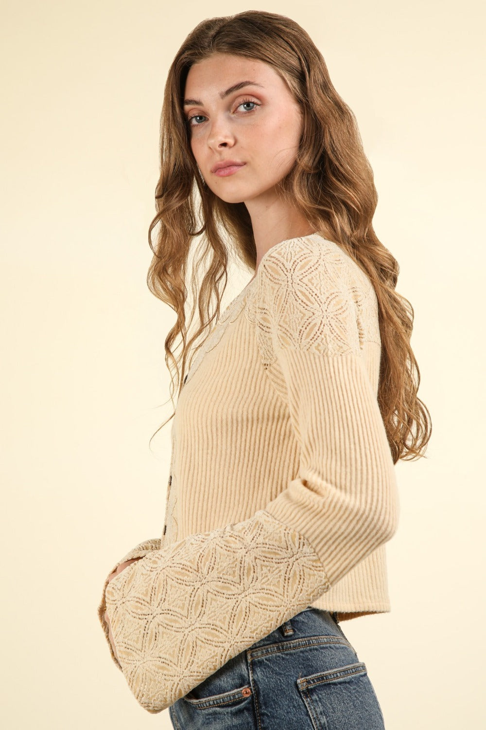 A woman with long brown hair is wearing the VERY J V-Neck Lace Detail Button Down Crop Ribbed Knit Top and blue jeans, standing against a plain background. The crop top features stylish bell sleeves that add a touch of elegance to her look.
