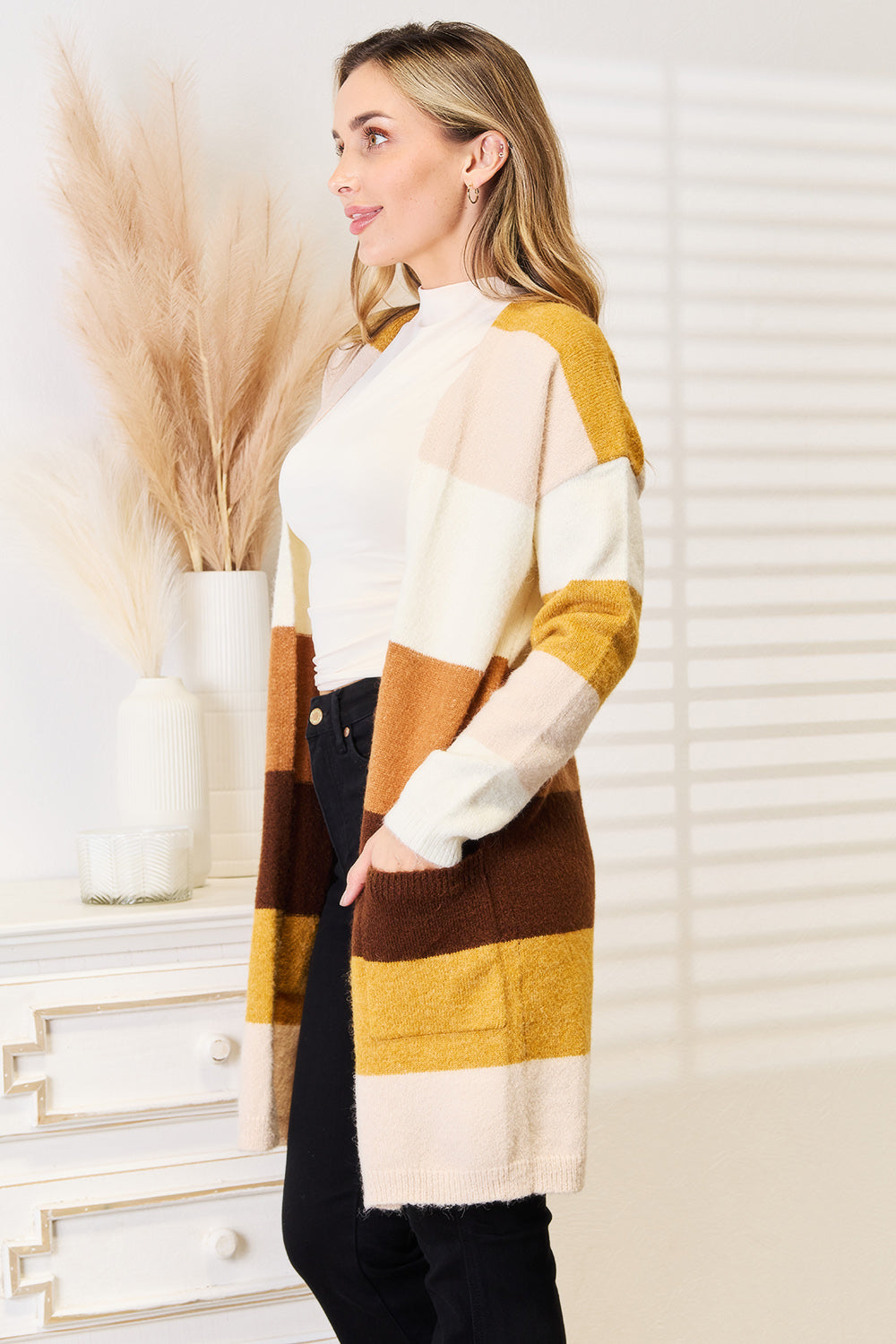 A woman wearing a cozy Woven Right Color Block Dropped Shoulder Cardigan, white turtleneck, and black pants stands indoors with her hands in her pockets.