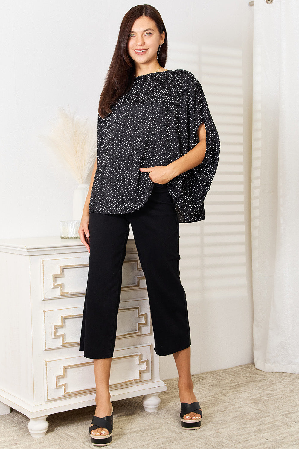 A woman stands wearing the Perfee Printed Dolman Sleeve Round Neck Blouse with white dots and black pants, smiling and looking to her left. The blouse features a printed design that adds a touch of elegance to her outfit.