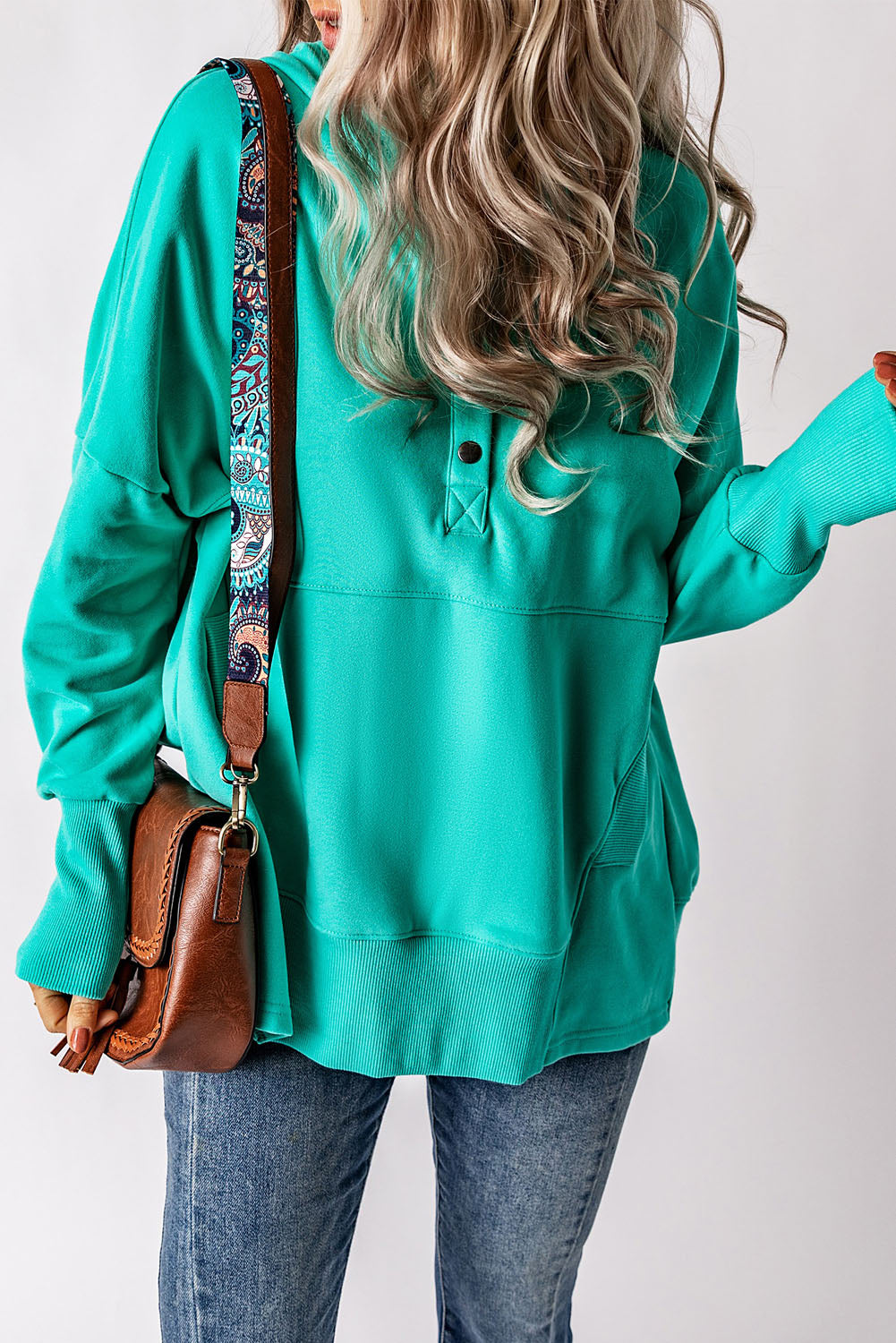 A person with long, wavy blonde hair is seen from the back wearing a Turquoise Batwing Sleeve Pocketed Henley Hoodie and carrying a brown bag with a patterned strap.