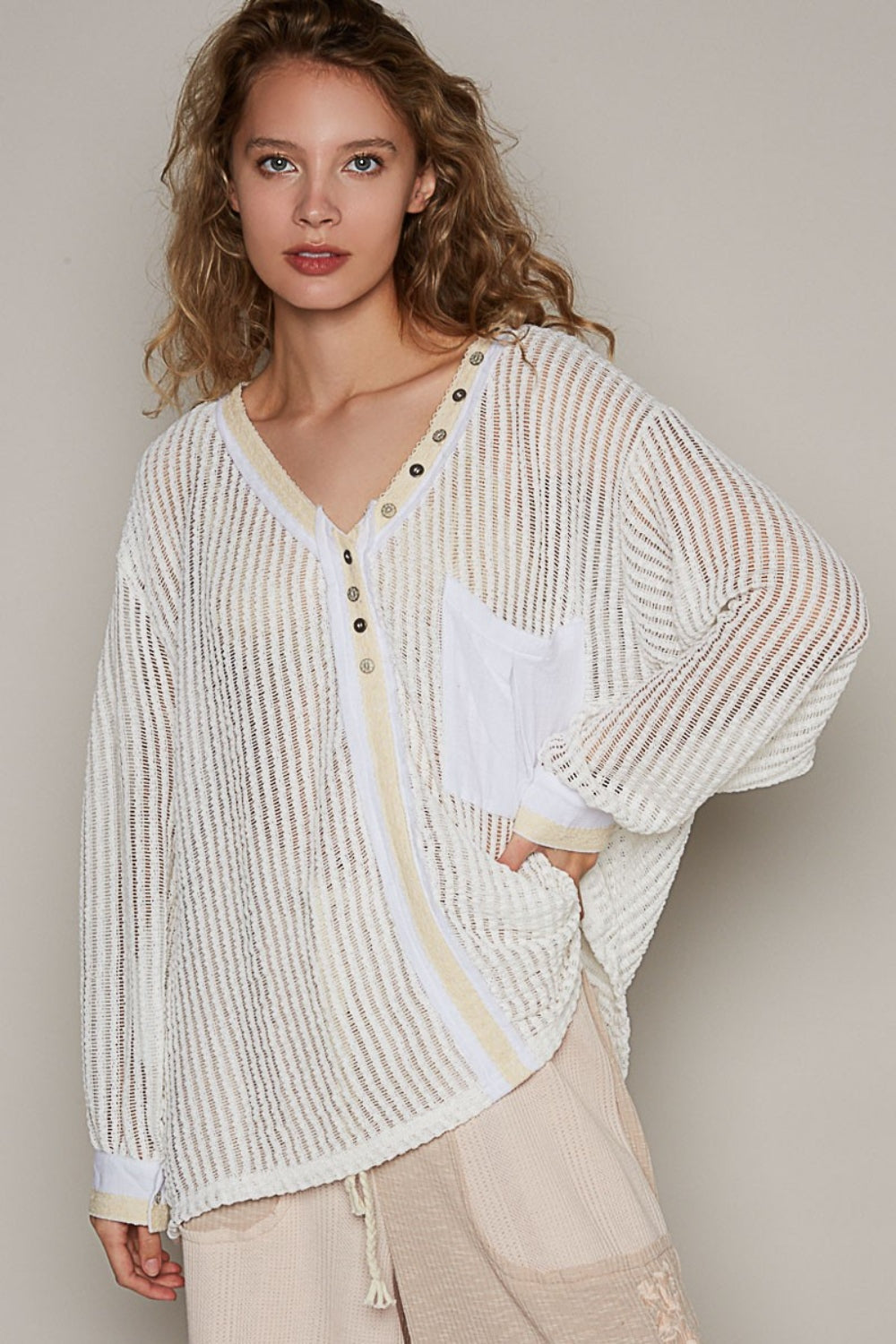 A person with wavy hair wearing a loose, bohemian-style POL V-Neck Long Sleeve Crochet Top in a sheer, striped white design paired with beige pants stands against a plain background.