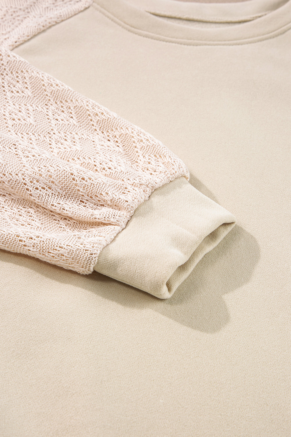 A blonde-haired individual wearing a Parchment Eyelet Knit Patchwork Raglan Sleeve Pullover Top stands indoors, smiling and looking to the side. Flowers and a ladder can be seen in the background.
