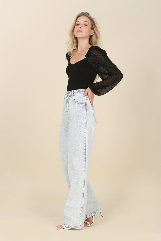 A person wearing the LS shirring sleeve bodysuit with Swiss dot mesh, featuring sheer, long puffed sleeves and a sweetheart neck, paired with light-colored jeans, stands against a plain background.