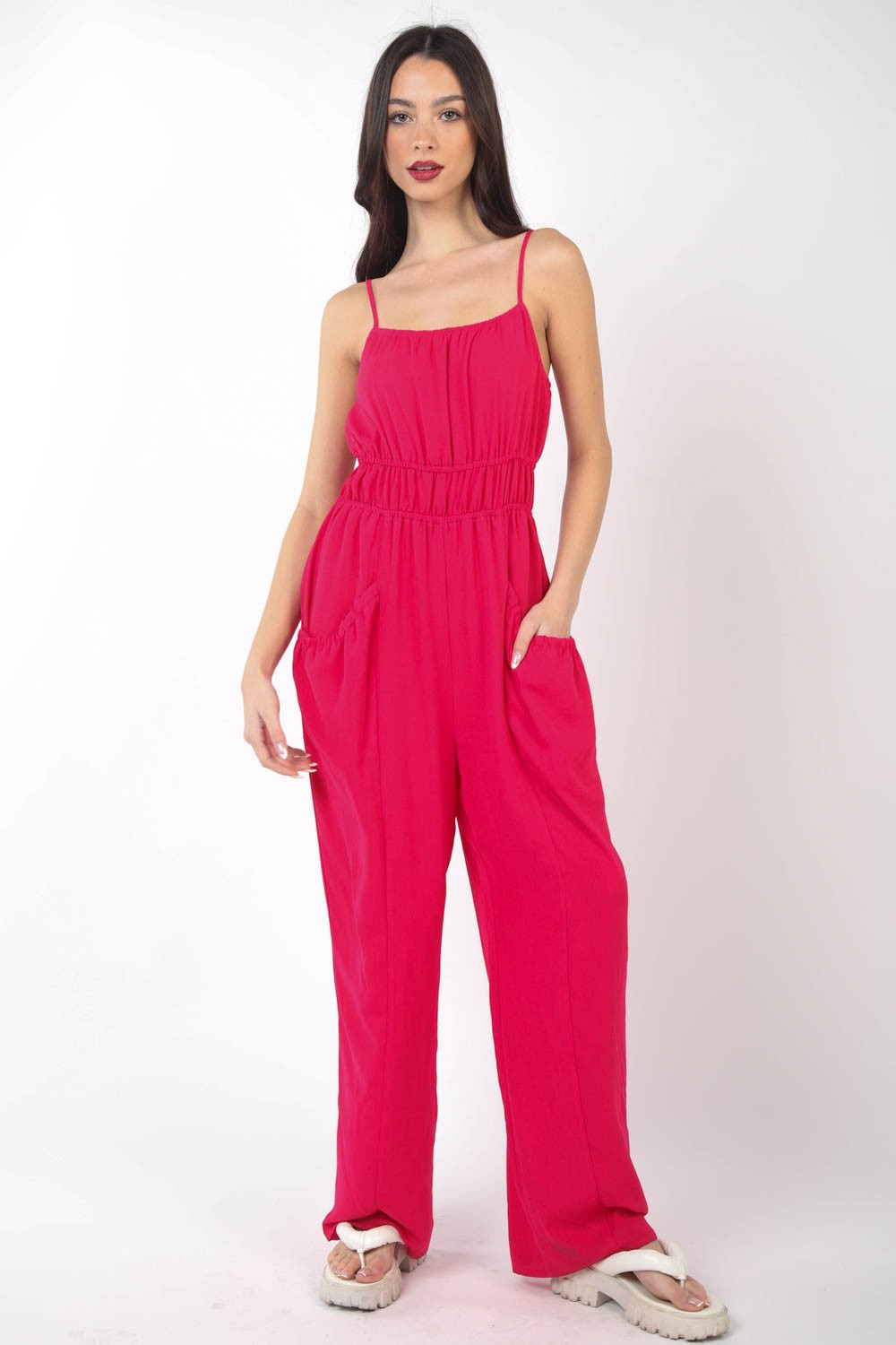 A person wearing the VERY J Pintuck Detail Woven Sleeveless Jumpsuit in bright pink paired with white sandals stands against a white background, with one hand placed in a pocket.