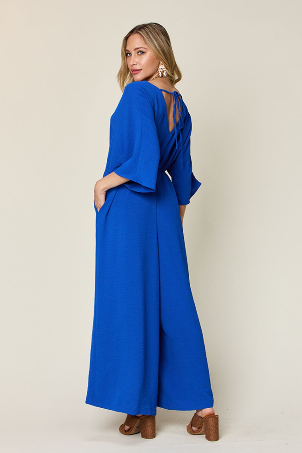 Person wearing the Double Take Full Size Surplice Wide Leg Jumpsuit with Pockets in royal blue, featuring wide legs, a deep V-neckline, and ruched detailing, posing with hands on hips against a plain light-colored background.