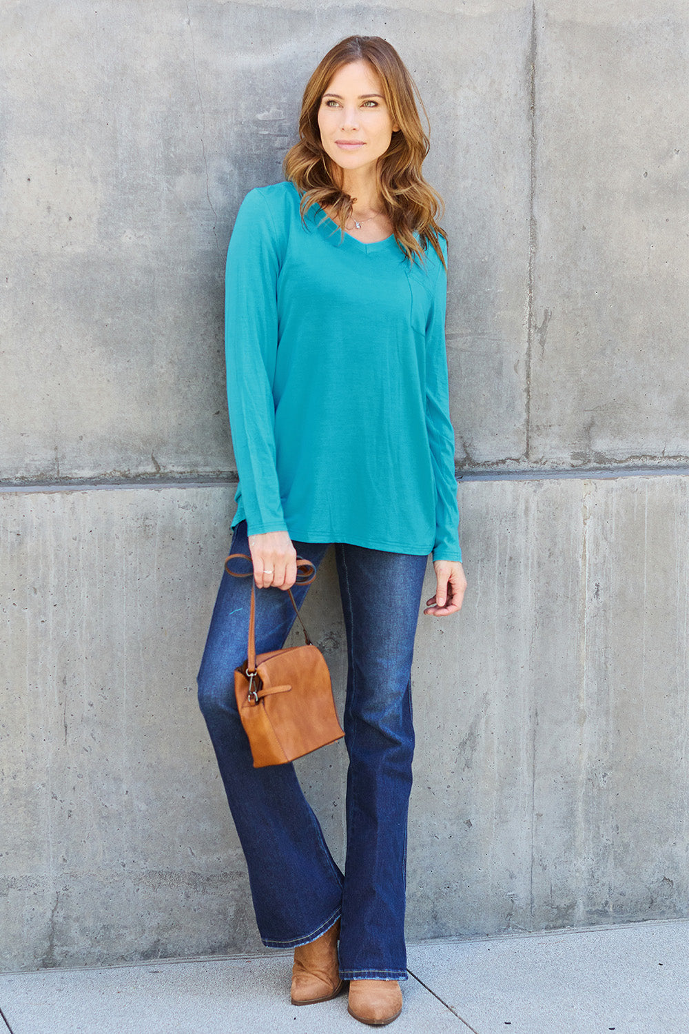 A woman with light brown hair stands against a concrete wall, wearing the Basic Bae Full Size V-Neck Long Sleeve Top in olive green and blue jeans, with a white backpack on her shoulder. Her outfit exudes basic style and is made from polyester, ensuring it’s both comfortable and suitable for machine wash cold. She is looking at the camera.