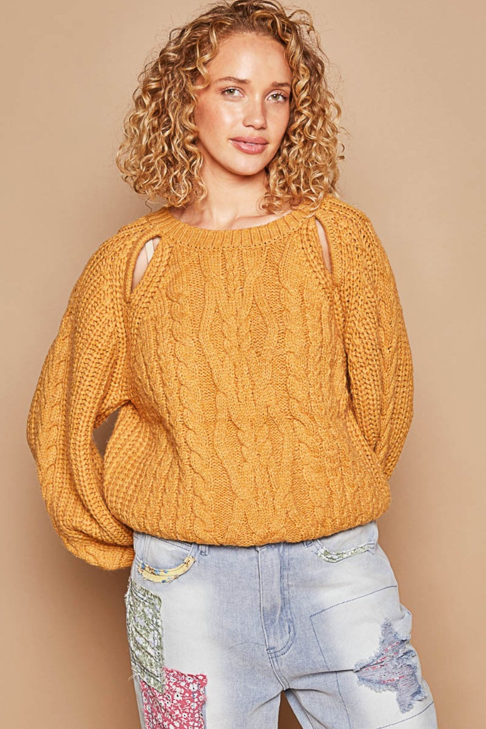 A person with curly hair wears a POL Cable Knit Cutout Long Sleeve Sweater in mustard, paired with light blue jeans adorned with patchwork designs.