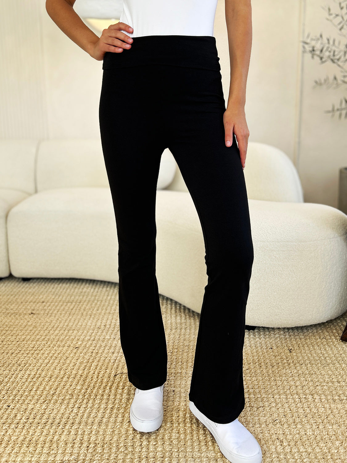 An individual dressed in Zenana Full Size High Waist Wide Waistband Bootcut Active Pants and white shoes, standing with a hand on the hip in front of a setting that includes a white couch and a plant.