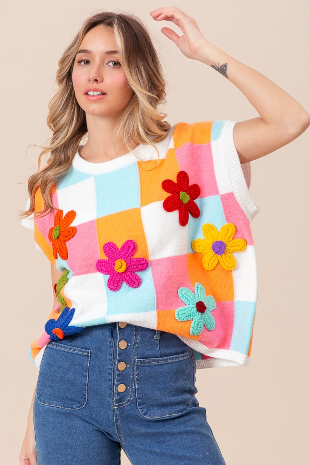 A woman is wearing the stylish BiBi Flower Patch Checkered Sweater Vest paired with high-waisted jeans. She has wavy blonde hair and a radiant smile while posing against a plain beige background. This trendy fall wardrobe piece adds a touch of warmth and whimsy to any outfit.