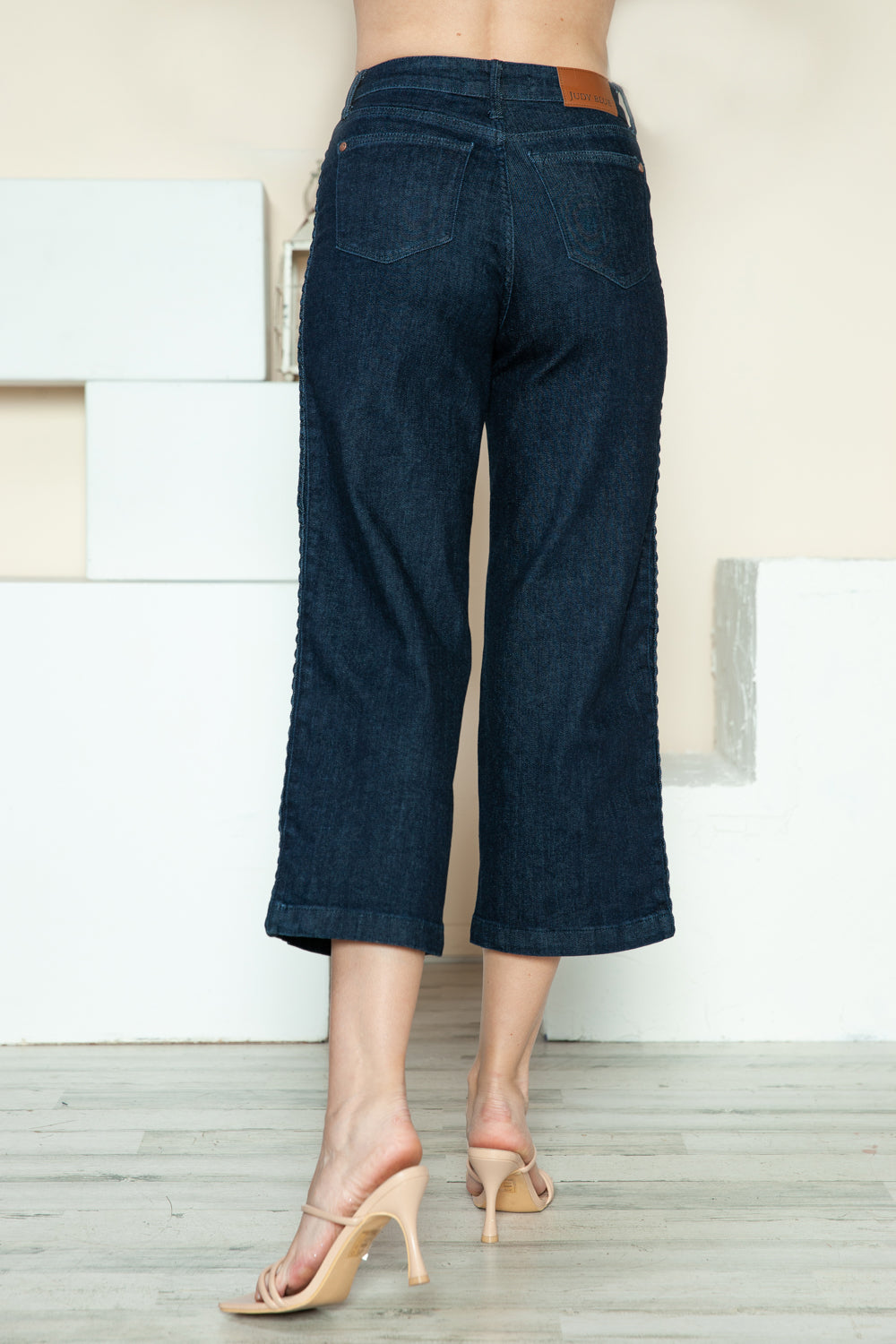 Person wearing Judy Blue Full Size Side Seam Braid Detail Crop Wide Leg Jeans and beige strappy heels, standing on a wooden floor with a white background.