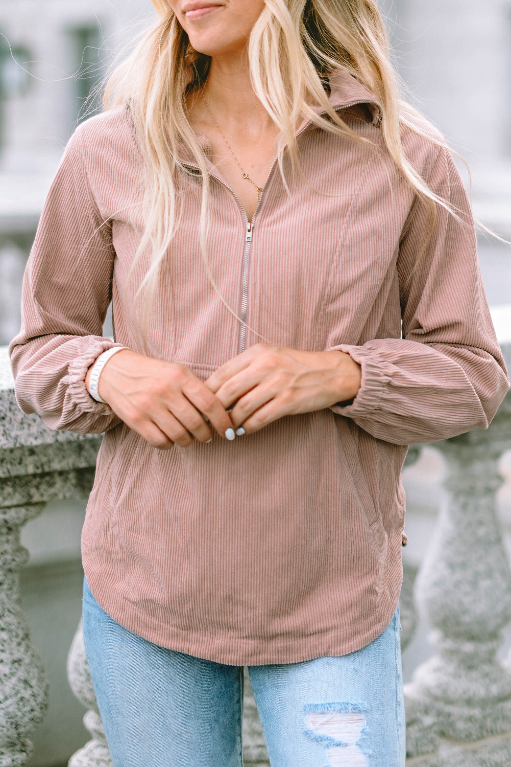 A person with long blonde hair, wearing a trendy Apricot Ribbed Texture Half Zip Collared Sweatshirt and blue jeans, stands facing away and looks over a stone balustrade.