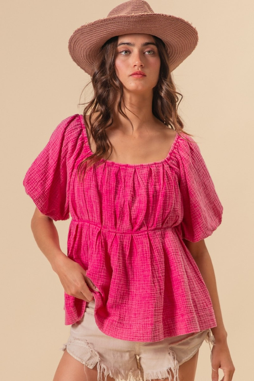 A woman wearing the BiBi Pleated Puff Sleeve Washed Blouse in pink, along with a wide-brimmed hat, strikes a pose against a beige background with one hand resting in her pocket.