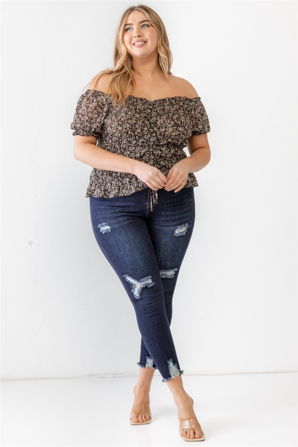 Wearing the Zenobia Plus Size Frill Ruched Off-Shoulder Short Sleeve Blouse and ripped jeans, a person with long hair poses against a plain white background.