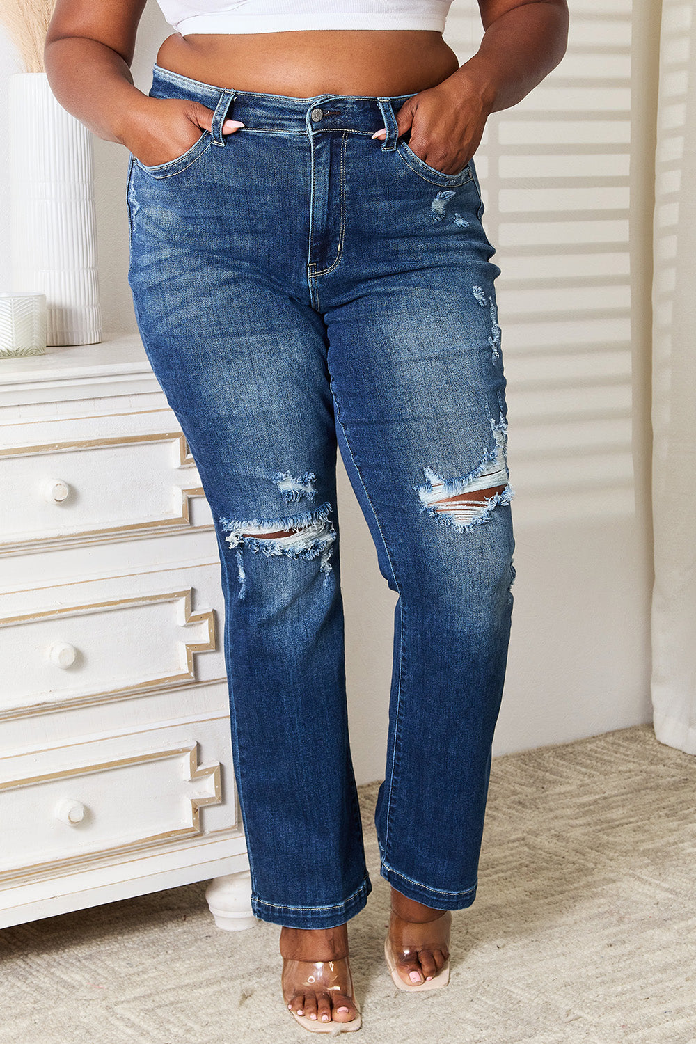 A person wearing Judy Blue Full Size Mid Rise Hand Sand & Destroy Bootcut Jeans stands with hands in pockets. The slightly stretchy jeans have rips on both legs, and the person is standing indoors beside a white dresser.