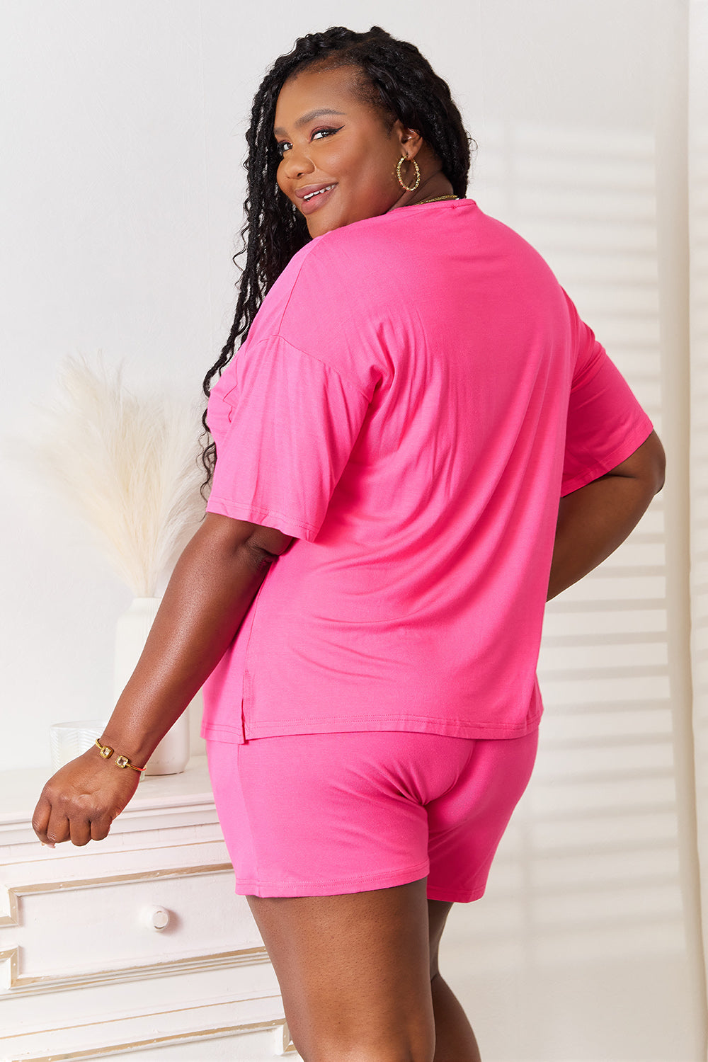 A woman is standing in a brightly lit room, wearing the Basic Bae Full Size Soft Rayon Half Sleeve Top and Shorts Set in a relaxed, stretchy turquoise fabric. She has one hand placed in her pocket and is smiling while looking slightly to the side.