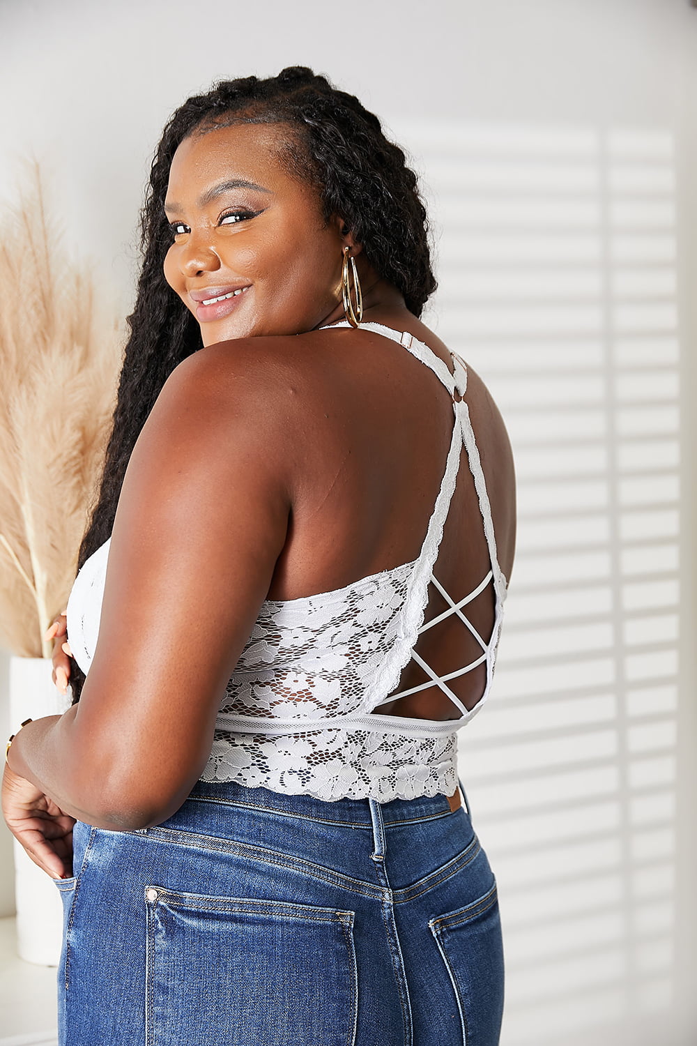 A woman gracefully stands in front of an indoor plant, wearing the JadyK Juliette Full Size Crisscross Lace Bralette paired with jeans.