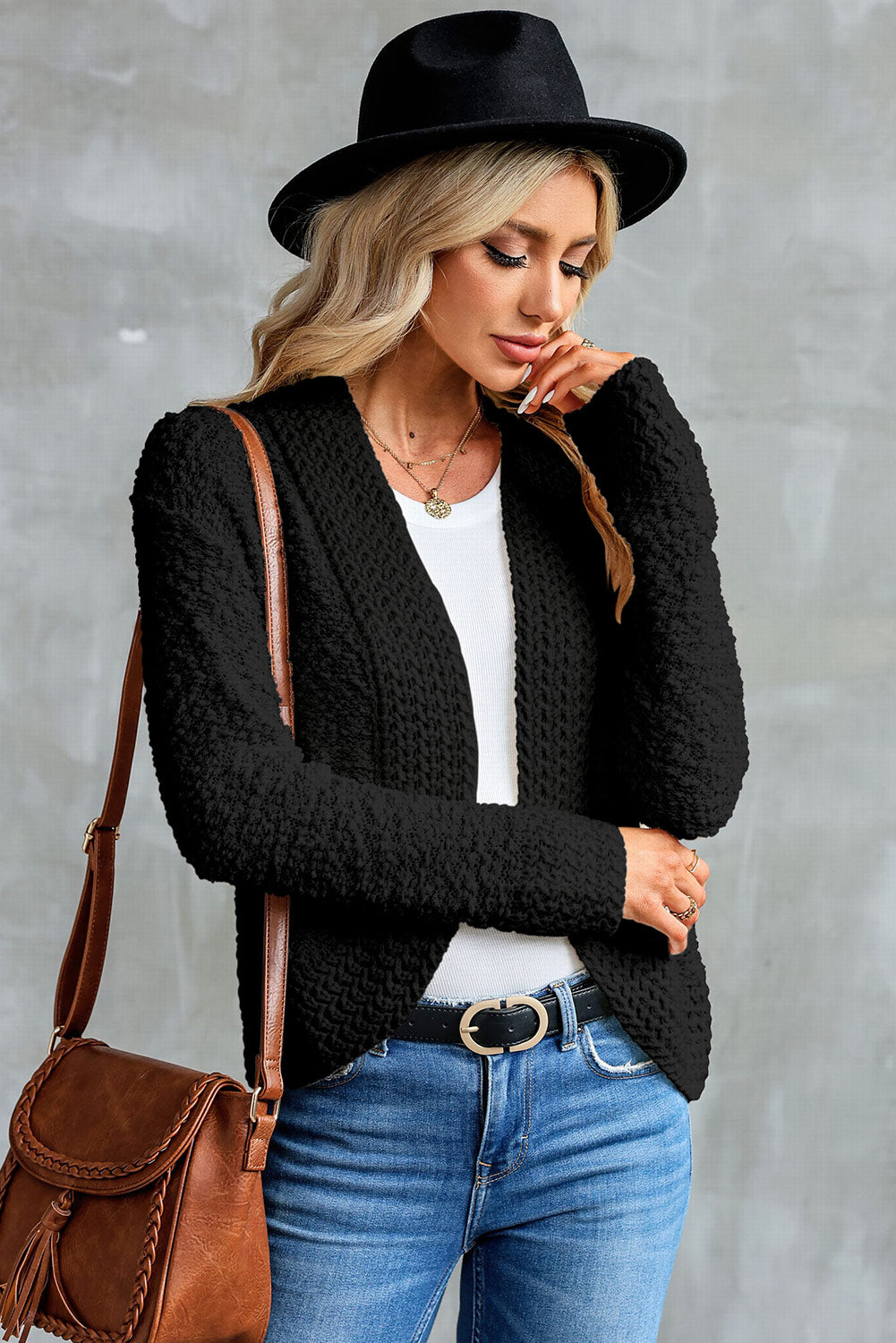 A woman in an Apricot Popcorn Knit Open Front Cardigan and jeans stands against a brick wall, looking to the side with a slight smile.