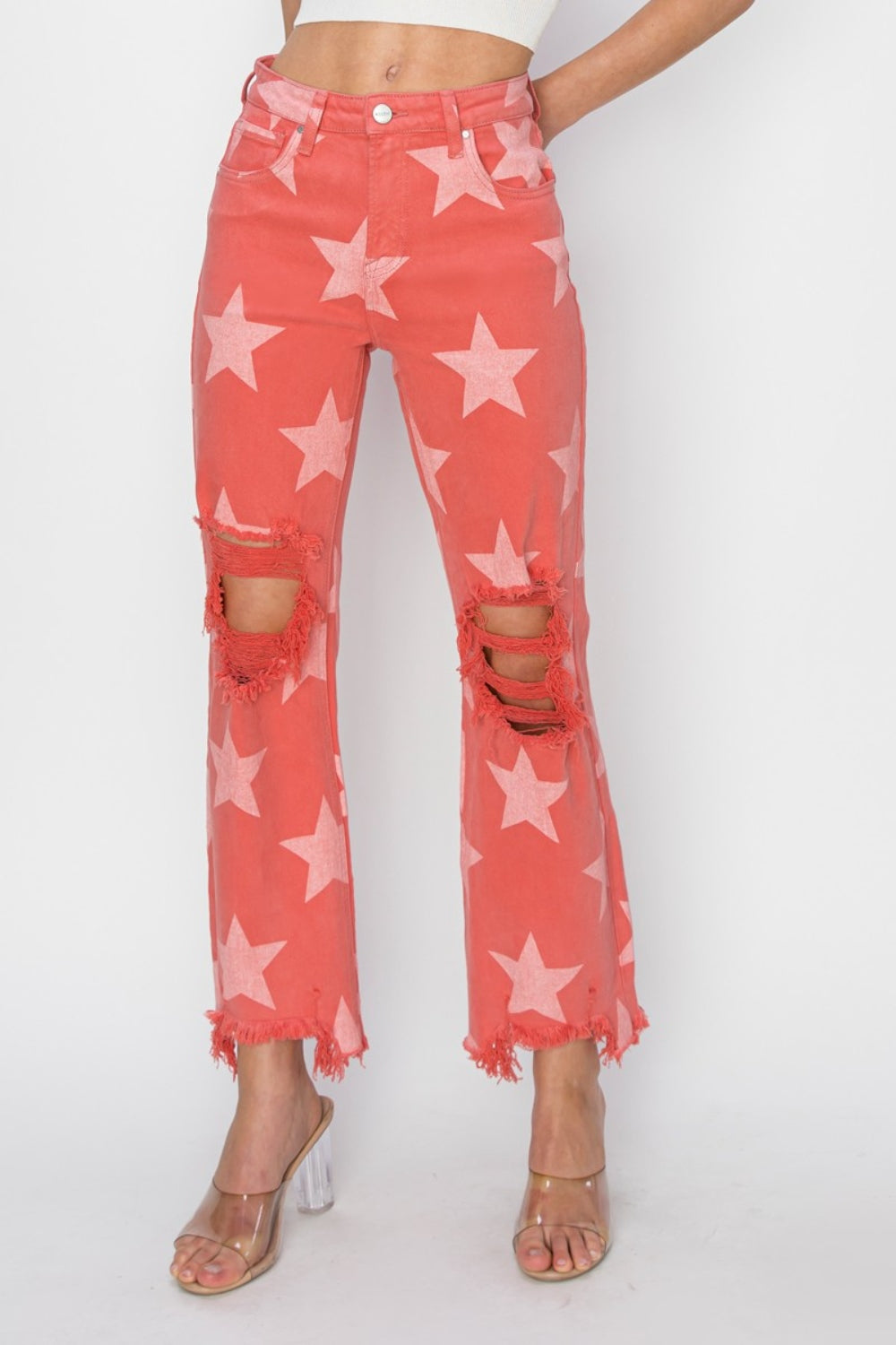 A person wearing the RISEN Full Size Distressed Raw Hem Star Pattern Jeans in a coral color, complete with distressed rips at the knees and raw frayed hems. The rock 'n' roll style is completed with clear high-heeled sandals.