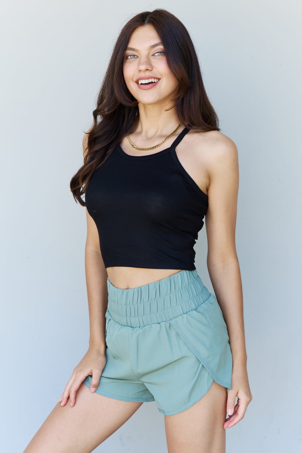 A person wearing a Ninexis Everyday Staple Soft Modal Short Strap Ribbed Tank Top in Black and light blue shorts is standing against a plain background, smiling slightly and looking to the side.