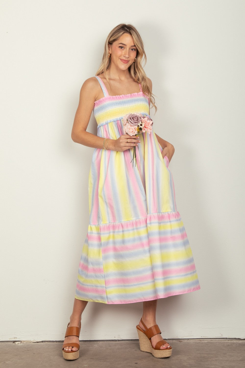 A woman in the VERY J Striped Woven Smocked Midi Cami Dress, featuring pastel stripes, holds a small bouquet of flowers. She pairs the dress with tan platform sandals that complete her chic summer look and stands against a plain white background.