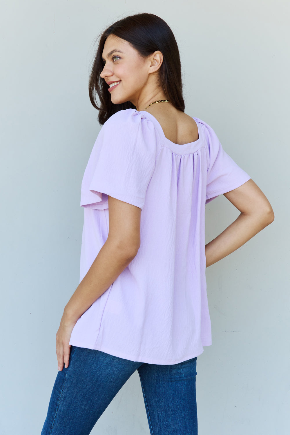 A person stands smiling in a lavender Ninexis Keep Me Close Square Neck Short Sleeve Blouse, paired with blue jeans against a plain background.