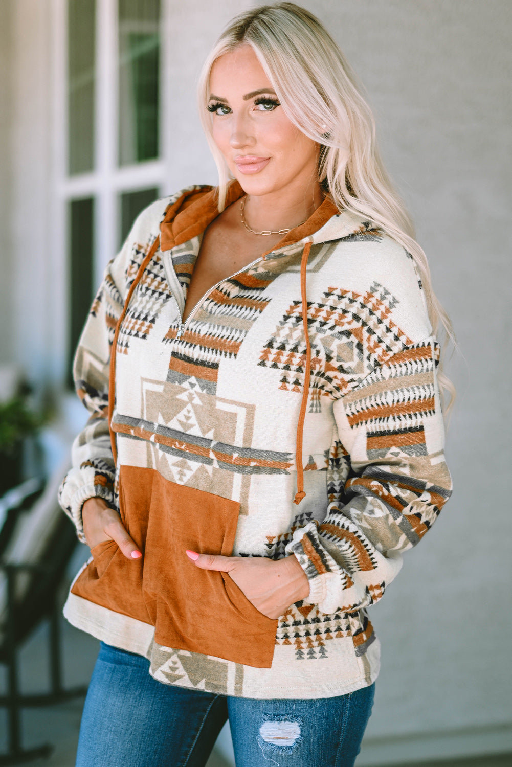 A person with long blonde hair, seen from behind, is wearing the Beige Aztec Print Kangaroo Pocket Half-Zip Hoodie. The hoodie showcases an Aztec pattern in shades of brown, beige, and orange and features a drop shoulder design with a cozy kangaroo pocket, all while standing outdoors.