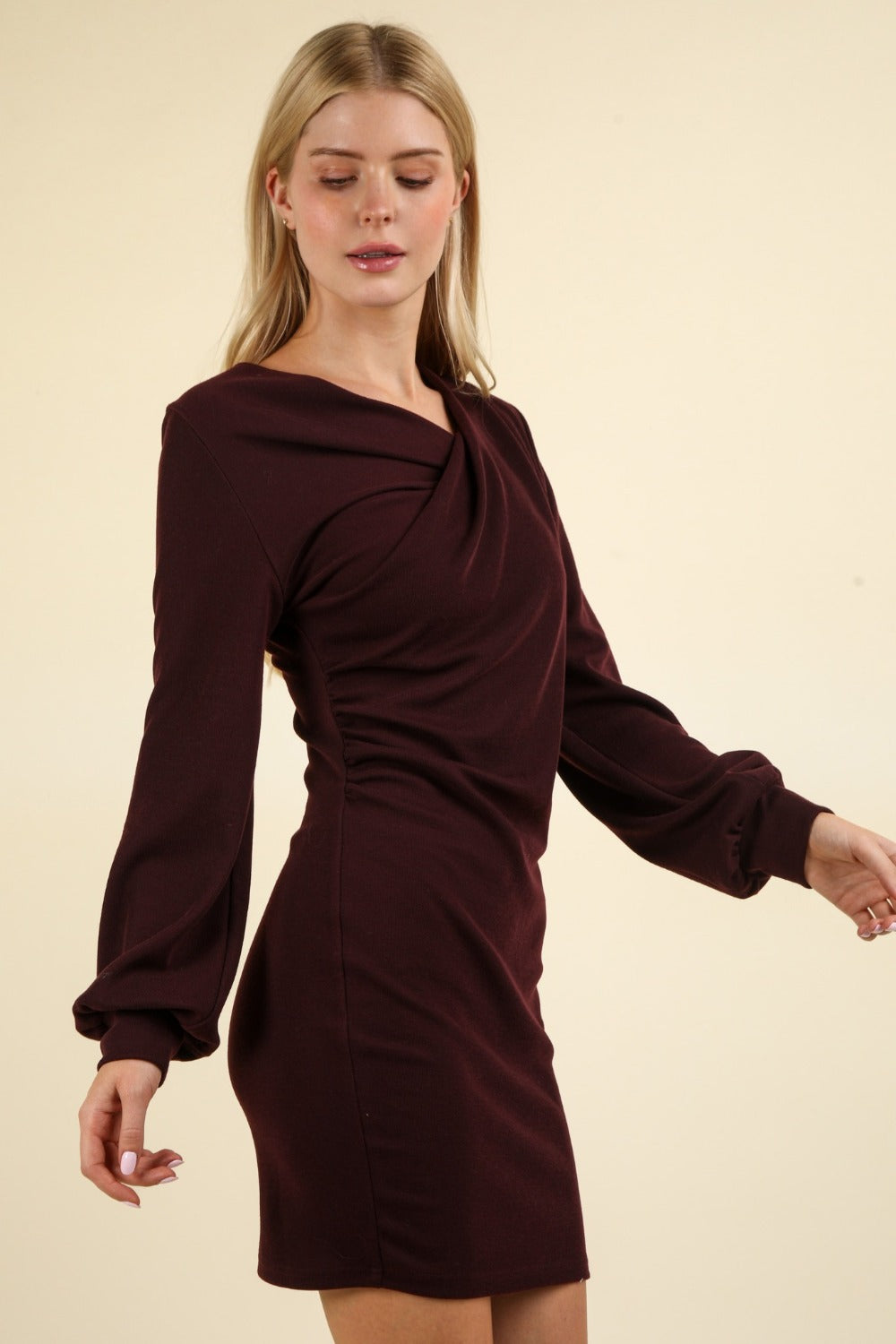 A person with long blonde hair is wearing a VERY J Ruched Detail Bodycon Mini Dress in burgundy, standing against a plain background. The figure-hugging silhouette of this dress makes it perfect for special occasions.