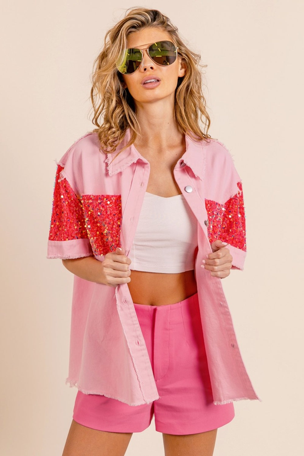 A woman poses in the BiBi Sequin Detail Raw Hem Short Sleeve Shirt, a pink button-up with red sequin detail pockets, paired with matching pink shorts and reflective aviator sunglasses.