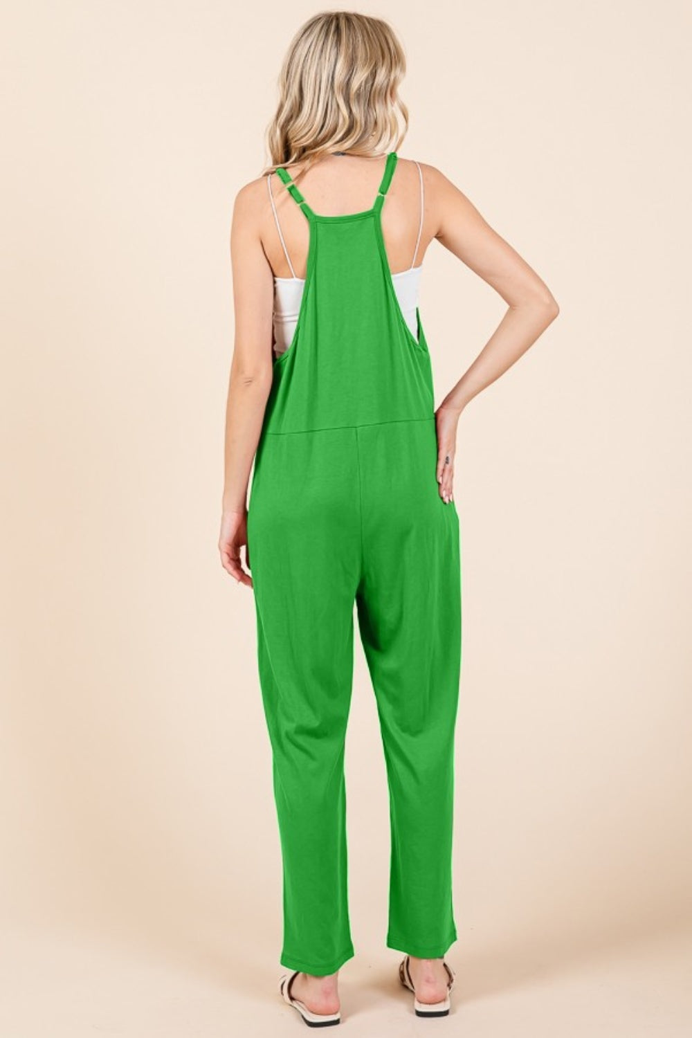 A person stands against a plain background wearing the Culture Code Full Size Sleeveless Jumpsuit with Pockets in green, paired with white sandals, showcasing its practical pockets with hands tucked comfortably inside.