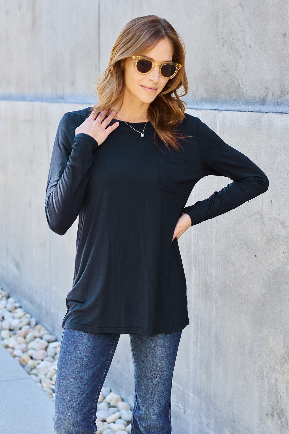 A woman with long hair wearing sunglasses, a Basic Bae Full Size Round Neck Long Sleeve Top in turquoise, and jeans is standing in front of a concrete wall with one hand on her hip.